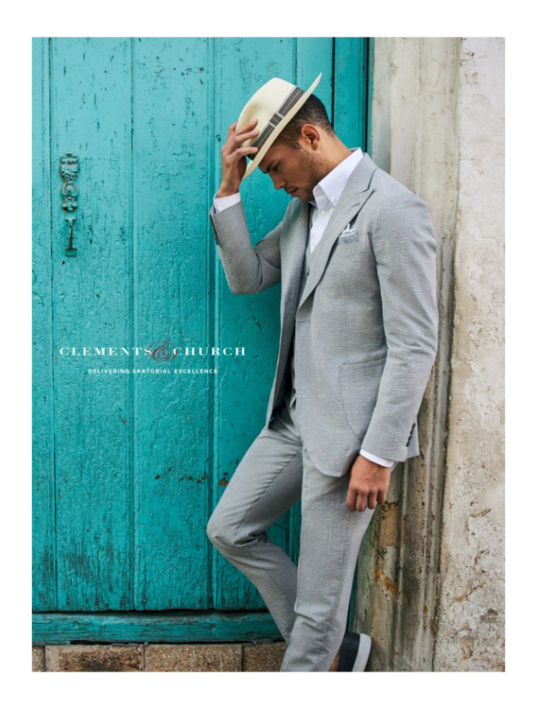 Man wearing a grey searsucker suit and tipping a Panama hat, leaning against a turquoise door in Havana, Cuba
