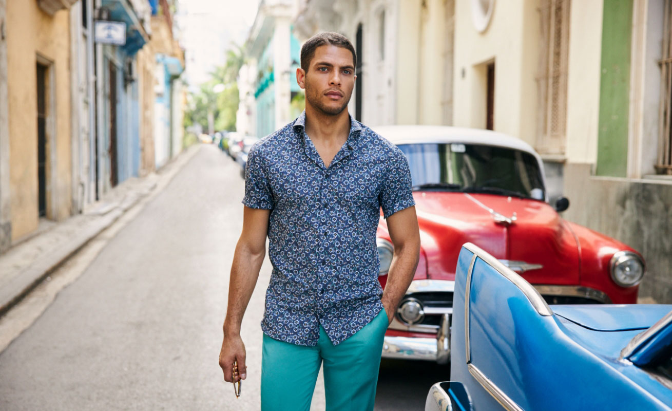 Man walking alongside red and blue vintage American cars, wearing a patterned blue shirt with bright turquoise trouseres, in a stree in Havana, Cuba